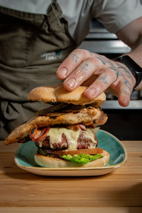 A chef finishing compiling a cheeseburger topped with a grilled chicken breast served on a bed of fresh lettuce and sliced tomato, styled on wooden boards.