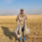 A person in a camouflage outfit and cap stands in a field, holding two large birds by their legs. The scene, epitomizing Nebraska waterfowl hunting, unfolds against a clear, blue sky and open grassy land.
