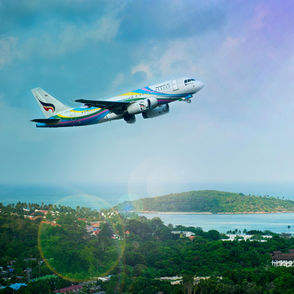 Commercial airplane taking off over tropical island coastline with lush greenery and ocean in the background, representing international air travel and vacation destinations.