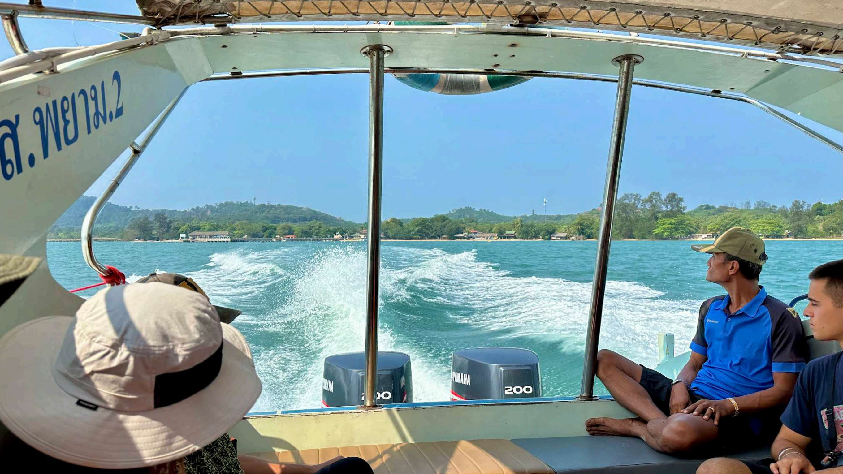Passengers aboard a covered boat looking across blue sea water towards a wooded island.