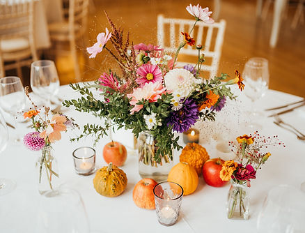 Colorful autumn wedding centerpiece with mixed wildflowers in a glass vase, surrounded by small pumpkins, apples, and lit votive candles on a white tablecloth in a rustic reception hall.