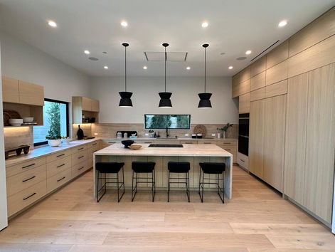Spacious and modern kitchen featuring light wood cabinetry with black handles, a large central island with a marble top and bar stools, set under black pendant lights. The kitchen is equipped with high-end appliances integrated into the cabinetry, creating a seamless and stylish look. Large windows provide ample natural light, enhancing the airy and inviting atmosphere of the space.