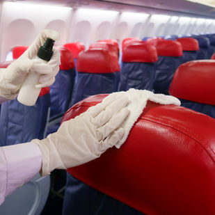 Gloved hands cleaning and disinfecting red leather aircraft seats with a spray bottle and cloth to ensure passenger hygiene and safety.
