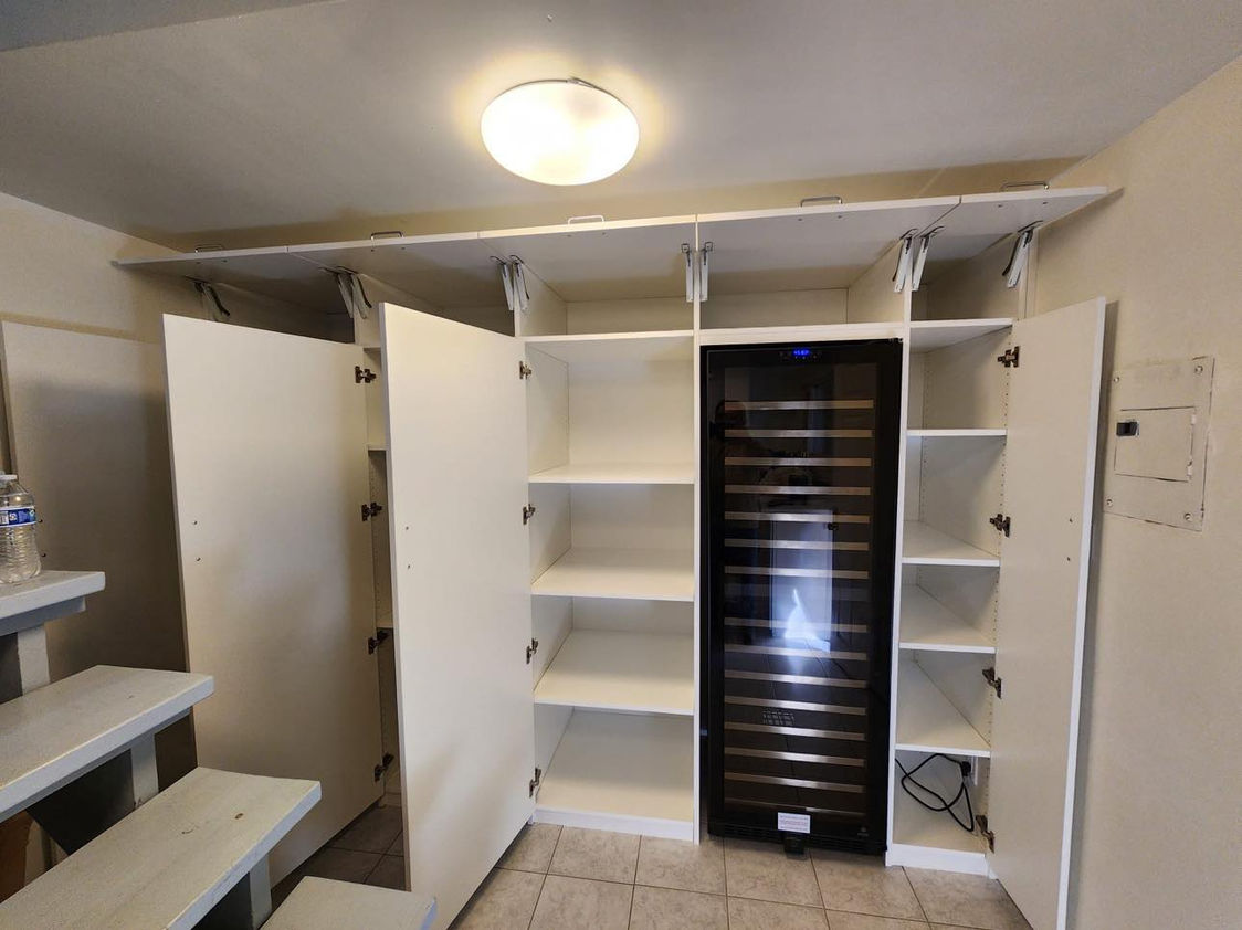 Functional utility room fitted with white storage cabinets, open shelving, and a central glass-door wine cooler. The doors are open, revealing ample shelving space ideal for household supplies or pantry items. The room is illuminated by a ceiling light, providing a bright and organized space for efficient storage and accessibility.