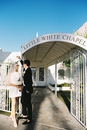 Bride and groom in sunglasses smile outside a white chapel under a clear blue sky. Veil and flowers; joyful wedding moment.