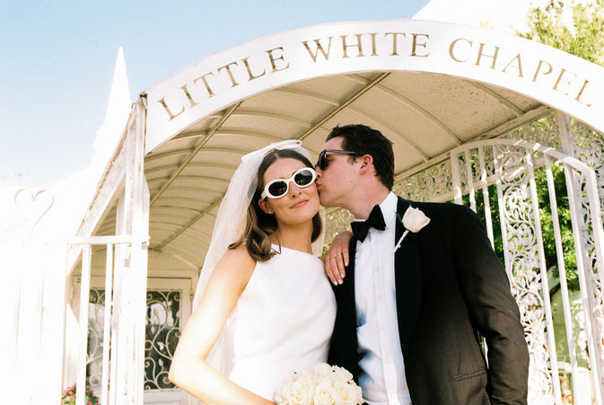 Bride and groom in sunglasses smile outside a white chapel under a clear blue sky. Veil and flowers; joyful wedding moment.