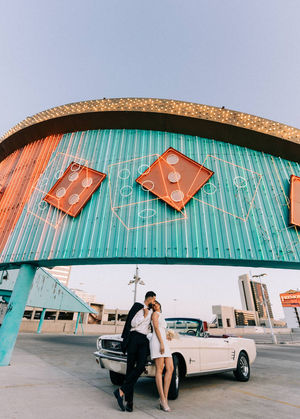 Bride and groom pose with classic convertible mustang under neon lights with teal and red dice. Where to take wedding pictures in Las Vegas. Captured by las Vegas elopement photographer brazen honey films.