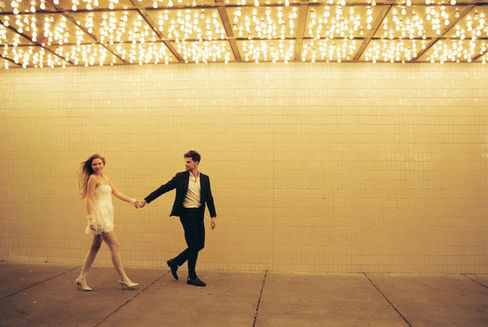 A couple in formal attire joyfully holds hands walking under glowing lights. Background: tiled wall, warm ambiance.