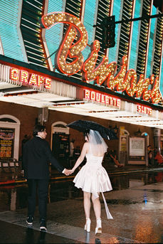 Couple under umbrella on wet street with neon hotel signs. Bride in white dress; groom in dark suit. Nighttime, city lights reflecting on pavement.