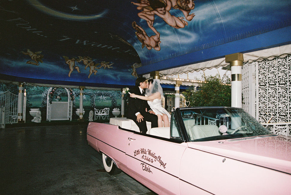Bride and groom joyfully kiss sitting inside a pink convertible at a wedding chapel. Cupid mural above. Bride wears a white dress, groom in a suit.