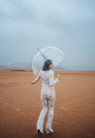 Bride holds a clear umbrella in the desert, wearing a white bridal jumpsuit on her elopement day. Captured by Las Vegas wedding photographer brazen honey films