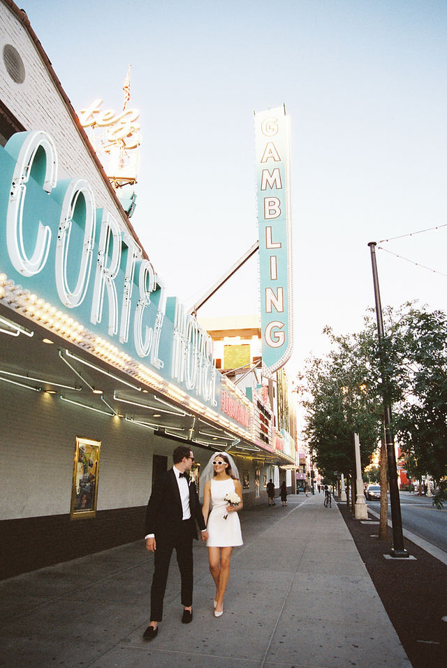 Bride and groom in sunglasses joyfully pose outside a lit-up venue. She holds white flowers; Lively, festive vibe.