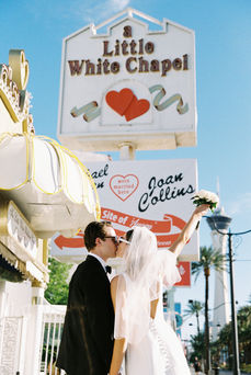 Bride and groom in sunglasses smile outside a white chapel under a clear blue sky. Veil and flowers; joyful wedding moment.