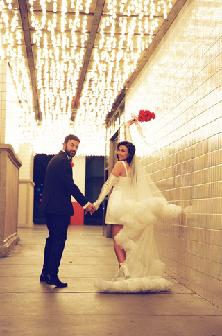 groom and bride wearing long veil with lots of tulle poses with red flowers underneath golden neon lights at the plaza hotel in las vegas