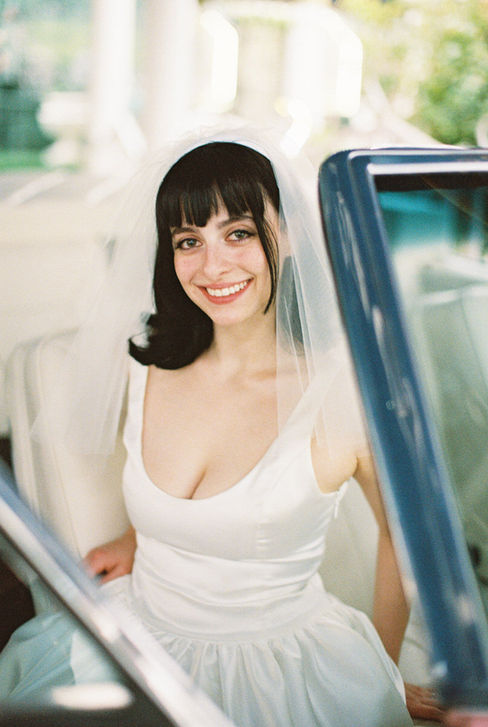bride with black hair and white dress sits in a convertible and smiles