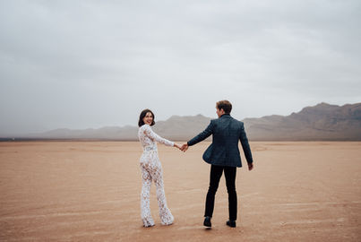 Bride & groom kiss underneath a clear umbrella in the desert on a rainy day. Bride wears a lace bridal jumpsuit, captured by Las Vegas wedding photographer brazen honey films