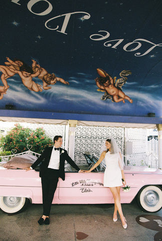 bride and groom hold hands and lean against the side of a pink convertible at the little white chapel with cupid mural above