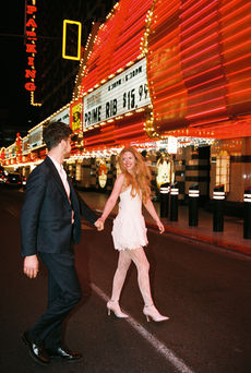 A couple in formal attire joyfully holds hands walking under glowing lights.