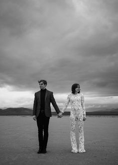 Moody black and white picture of bride and groom holding hands on their elopement out in the desert during a storm. Captured by Las Vegas elopement photographer brazen honey films. Bride wears a bridal jumpsuit
