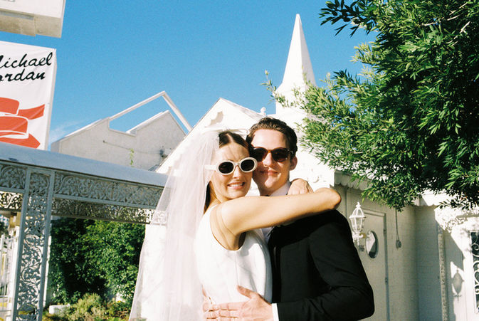 Bride and groom in sunglasses smile outside a white chapel under a clear blue sky. Veil and flowers; joyful wedding moment.