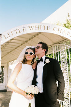 Bride and groom in sunglasses smile outside a white chapel under a clear blue sky. Veil and flowers; joyful wedding moment.