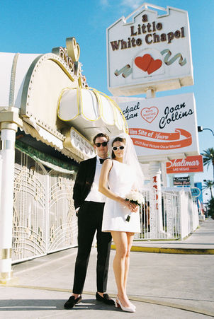 Bride and groom in sunglasses smile outside a white chapel under a clear blue sky. Veil and flowers; joyful wedding moment.