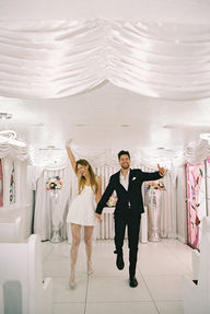 Bride in a white dress dances joyfully, groom claps, performer in gold jacket sings. Elegant floral backdrop with white drapery.