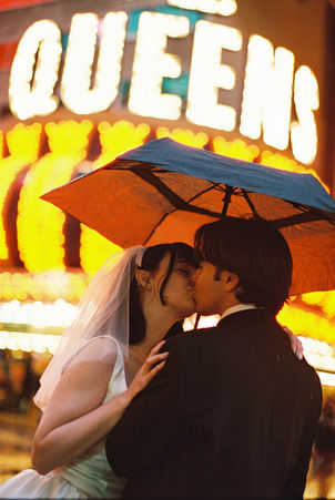 Couple under umbrella on wet street with neon hotel signs. Bride in white dress; groom in dark suit. Nighttime, city lights reflecting on pavement. kissing underneath an umbrella