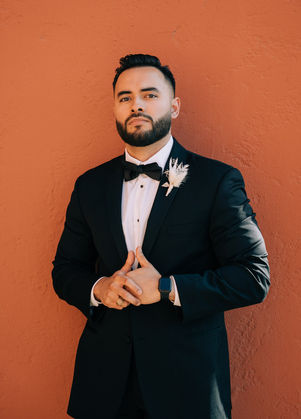 Groom wears tuxedo posing in front of a historic orange building in downtown Las Vegas. Best places to take wedding day pictures in Las Vegas. captured by las Vegas elopement photographer brazen honey films