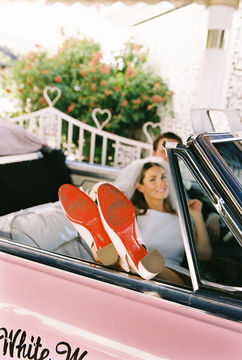 bride wearing red bottom shoes sits in pink car with feet out the window