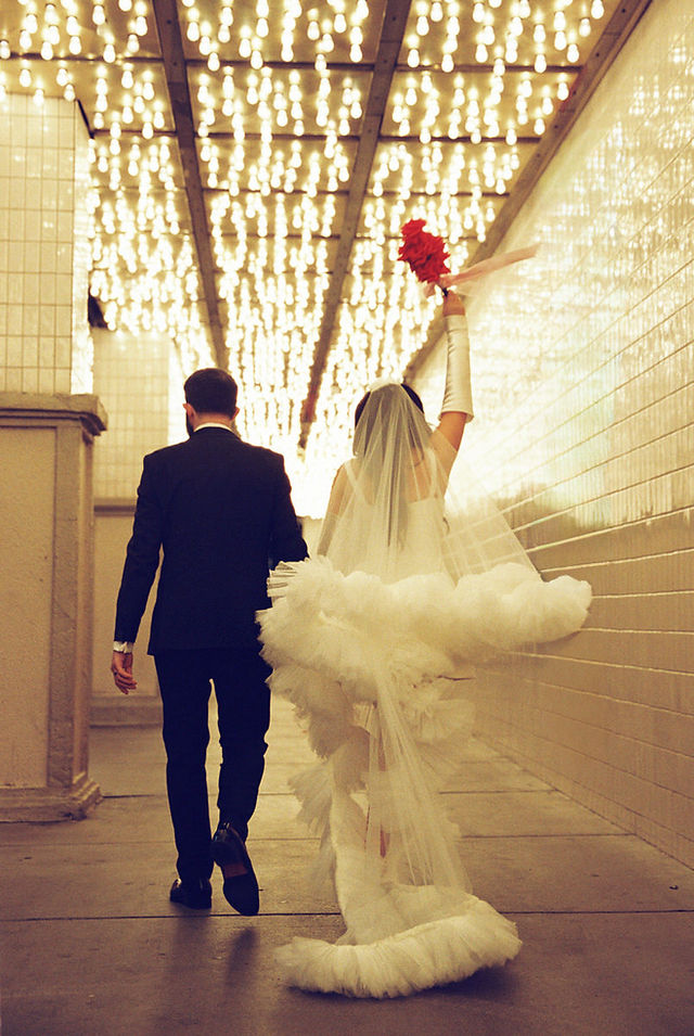 groom and bride wearing long veil with lots of tulle poses with red flowers underneath golden neon lights at the plaza hotel in las vegas