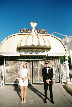 Bride and groom in sunglasses smile outside a white chapel under a clear blue sky. Veil and flowers; joyful wedding moment.