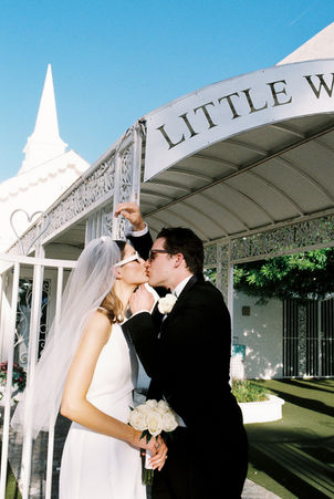 Bride and groom in sunglasses smile outside a white chapel under a clear blue sky. Veil and flowers; joyful wedding moment.