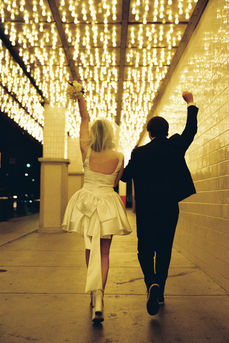bride wearing white dress embraces groom while holding flowers and smiling underneath golden neon lights