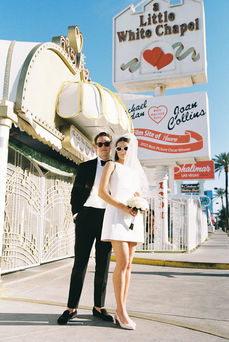 Bride and groom in sunglasses smile outside a white chapel under a clear blue sky. Veil and flowers; joyful wedding moment.