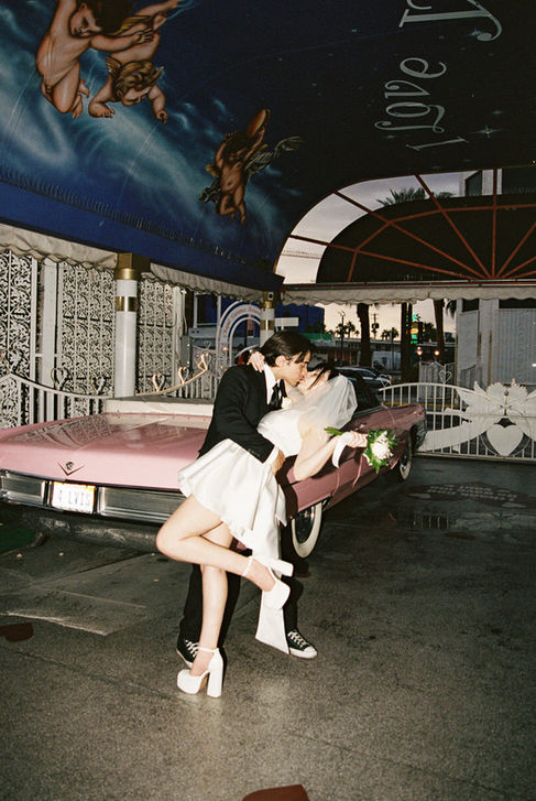Bride and groom joyfully raise arms next to a pink convertible at a wedding chapel. Cupid mural above. Bride wears a white dress, groom in a suit.