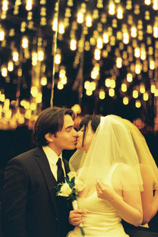 bride wearing white dress embraces groom while holding flowers and smiling underneath golden neon lights