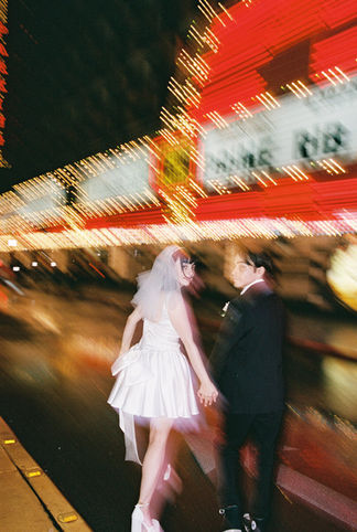 Couple under umbrella on wet street with neon hotel signs. Bride in white dress; groom in dark suit. Nighttime, city lights reflecting on pavement.