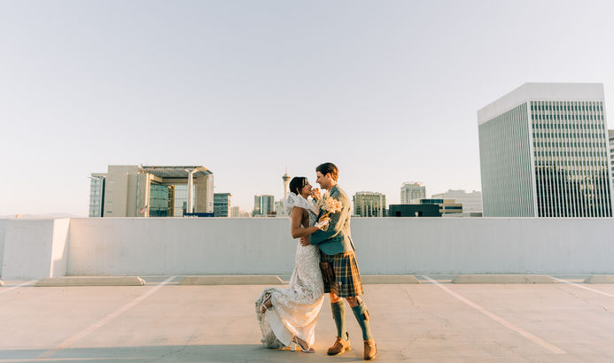 Bride and groom pose for pictures in downtown las vegas at golden hour with the strip in the background. captured by las vegas wedding photographer brazen honey films. Groom wears stylish stottish kilt. Bride wears custom made with love bridal gown.