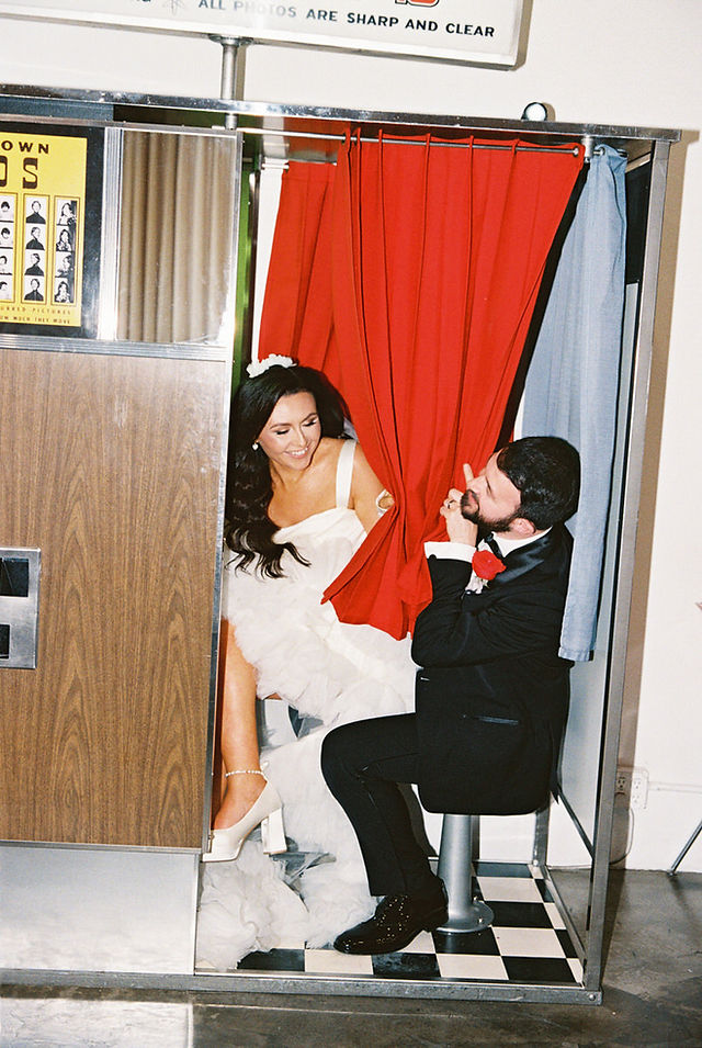 couple wearing wedding attire take photos in a analog photo booth behind a red curtain