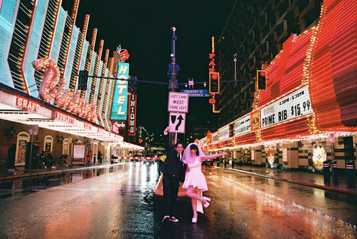 Couple under umbrella on wet street with neon hotel signs. Bride in white dress; groom in dark suit. Nighttime, city lights reflecting on pavement.