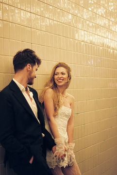 A couple in formal attire joyfully holds hands walking under glowing lights. Background: tiled wall, warm ambiance.