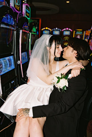 bride wearing white sits on groom lap in front of slot machines and kiss