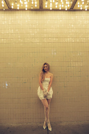 A couple in formal attire joyfully holds hands walking under glowing lights. Background: tiled wall, warm ambiance.