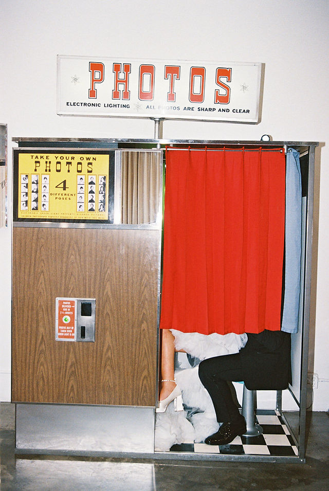 couple wearing wedding attire take photos in a analog photo booth behind a red curtain