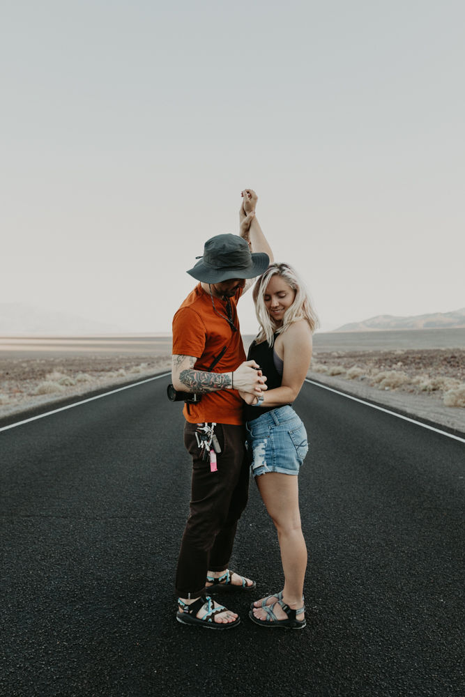 man and woman holding hands in the middle of a long and straight black road in the desert in death valley national park, captured by las vegas elopement photographer hayway films