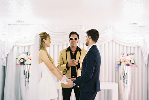 Bride in a white dress dances joyfully, groom claps, performer in gold jacket sings. Elegant floral backdrop with white drapery.