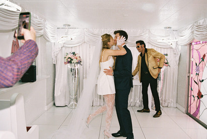 Bride in a white dress dances joyfully, groom claps, performer in gold jacket sings. Elegant floral backdrop with white drapery.