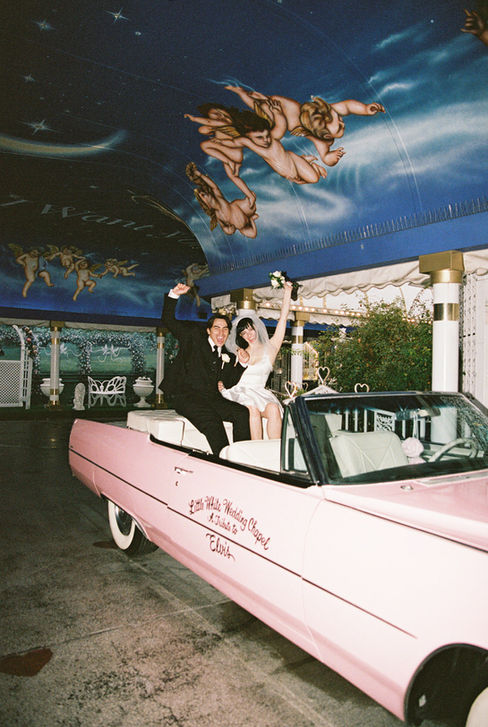 Bride and groom joyfully raise arms next to a pink convertible at a wedding chapel. Cupid mural above. Bride wears a white dress, groom in a suit.