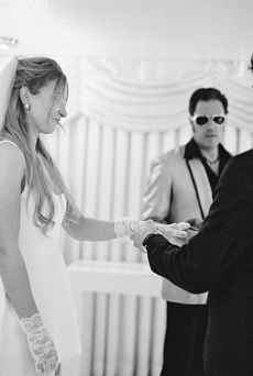 Bride in a white dress dances joyfully, groom claps, performer in gold jacket sings. Elegant floral backdrop with white drapery.